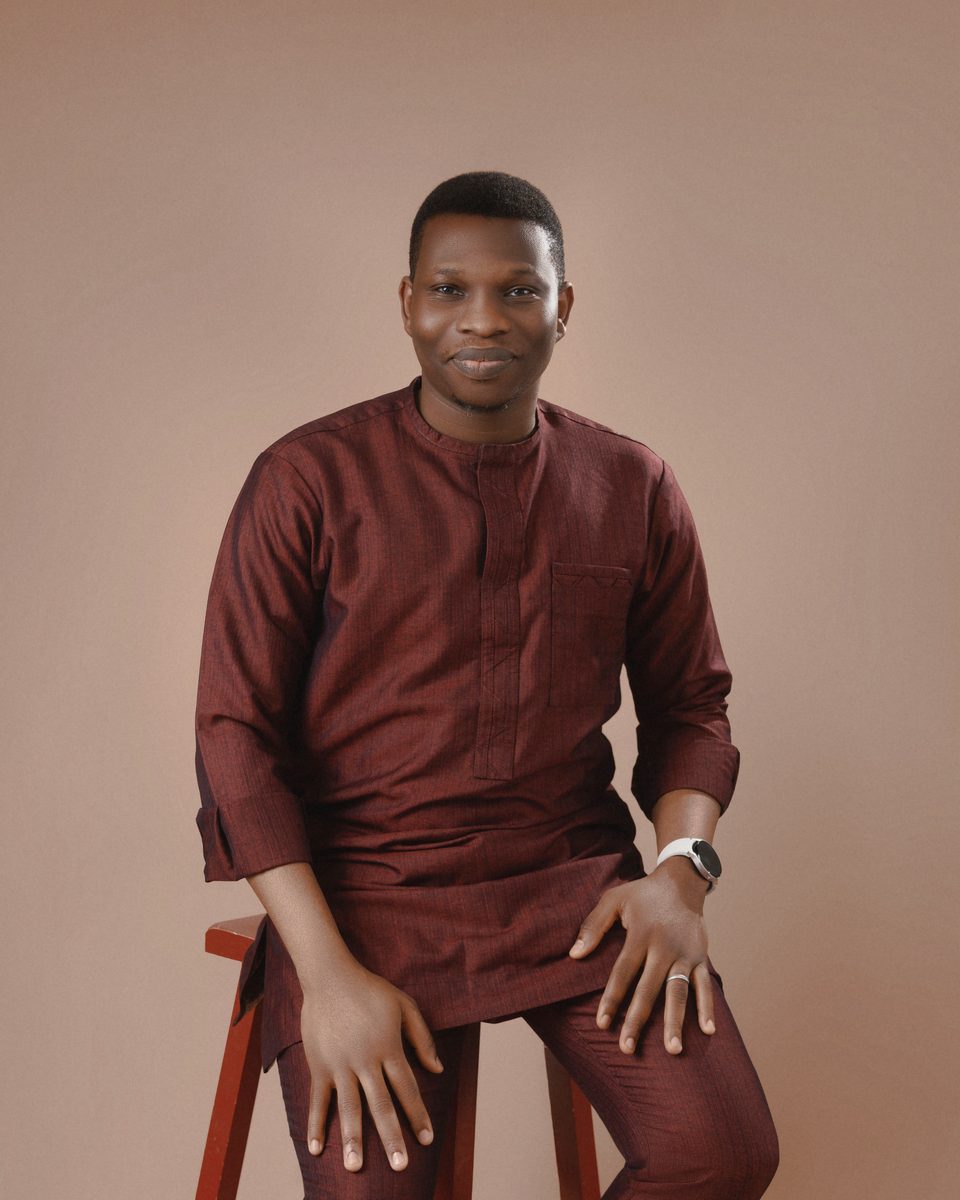 Victor Bolu in burgundy Nigerian attire, seated on a wooden stool and smiling