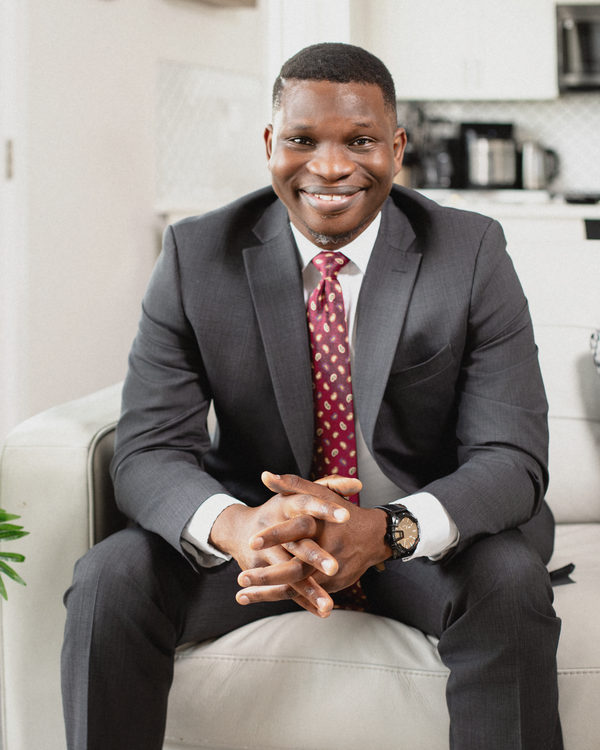 Victor Bolu in a charcoal suit and burgundy tie, seated on a sofa at home and smiling