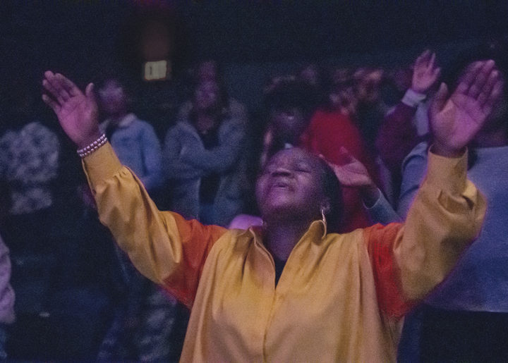 A woman in a yellow jacket lifts both arms high, head back and eyes closed, the room behind her with hands raised