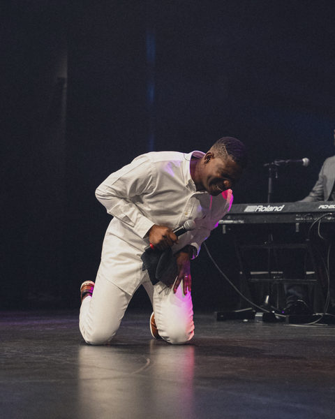 Victor kneeling on the stage floor in white, head bowed, microphone in hand