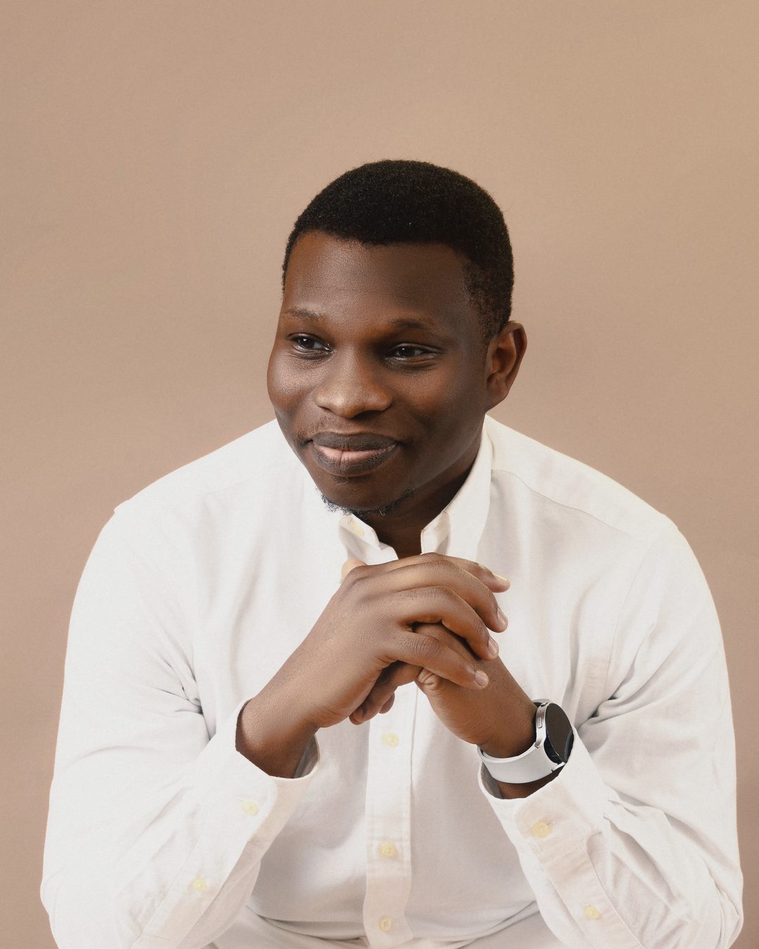 Victor Bolu in white linen, seated with his hands clasped, smiling as he looks to one side